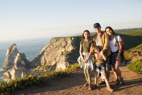 Happy father hiking with children in mountains. Portrait of mid adult man posing with teen daughters and preteen son against beautiful seascape. Active family weekend concept